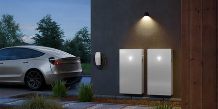 Two Tesla Powerwall units installed on a home’s exterior wall, with a Tesla electric car charging nearby. The Tesla Battery stores solar energy, providing backup power and reducing reliance on the grid.